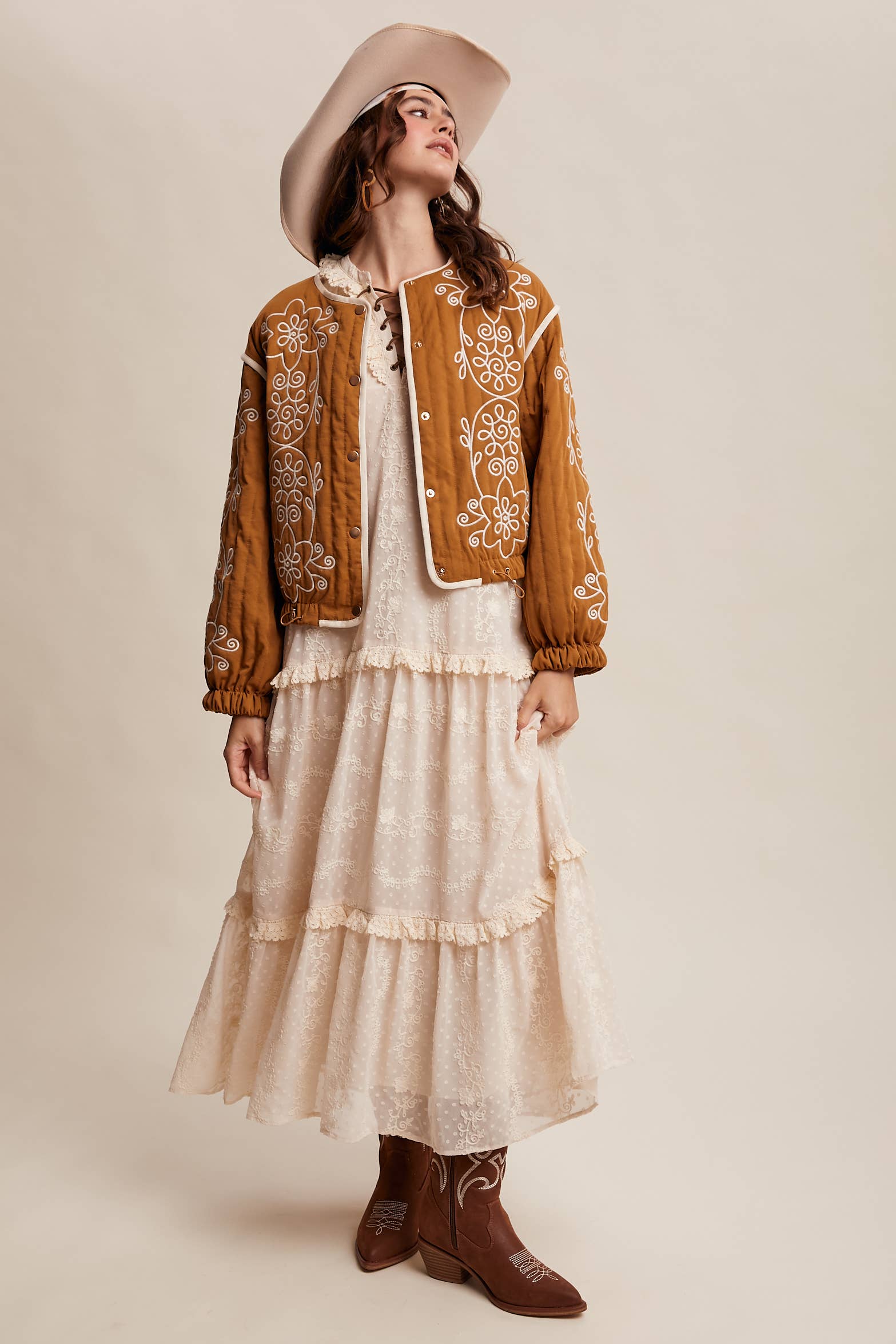 Boho caramel-brown jacket with cream floral swirl embroidery, ivory piping, and snap buttons, styled over a lace tie-front top with a scarf and felt hat outdoors.