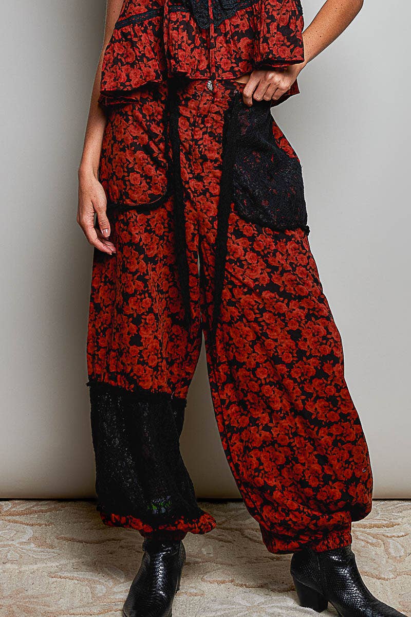 A person wearing loose, balloon-style pants with a red and black floral pattern and black lace trim, paired with black ankle boots, standing against a plain light wall.