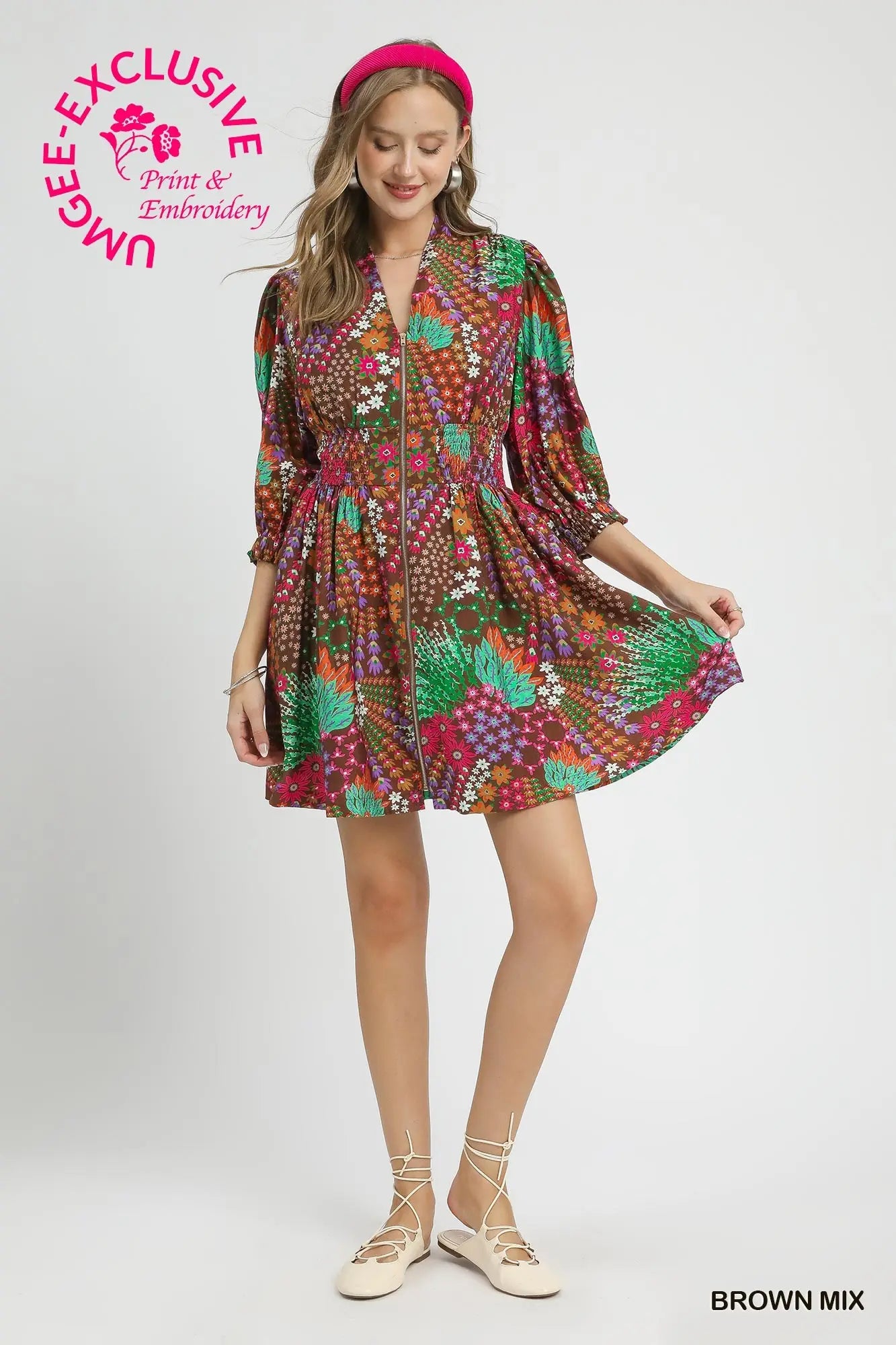 A woman modeling a short, knee-length dress with a dense multicolor floral and geometric print in red, green, orange, and brown tones. She is wearing a pink headband and beige lace-up flats against a plain light gray studio background.