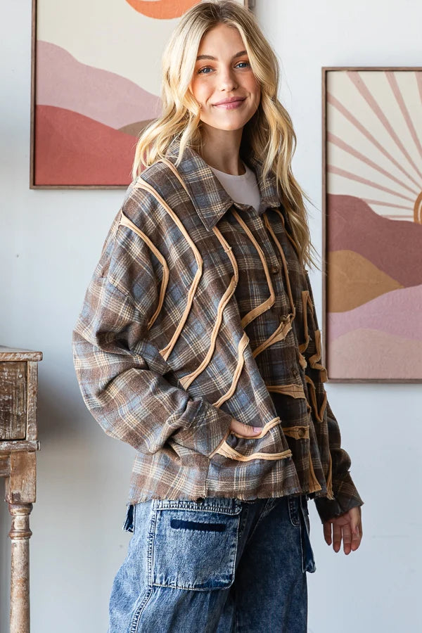 A woman wearing a brown and beige plaid button-up shirt jacket with diagonal tan stripe details, paired with blue jeans, standing in front of a wall with framed art.
