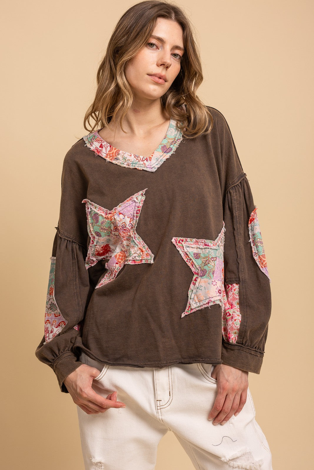 A woman wearing a brown long-sleeve top with floral star patches and a floral collar, paired with white pants, against a solid beige background.