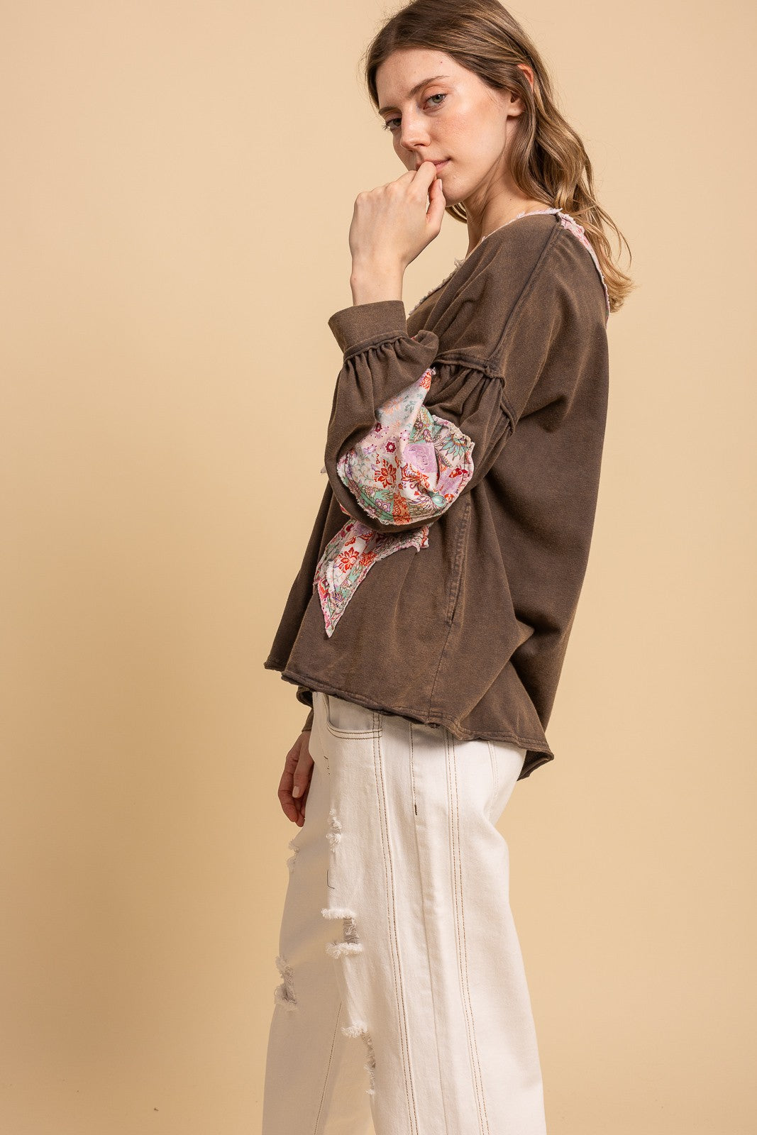 A woman wearing a brown long-sleeve top with a floral patterned underarm pouch and white pants, posed against a solid beige studio background.