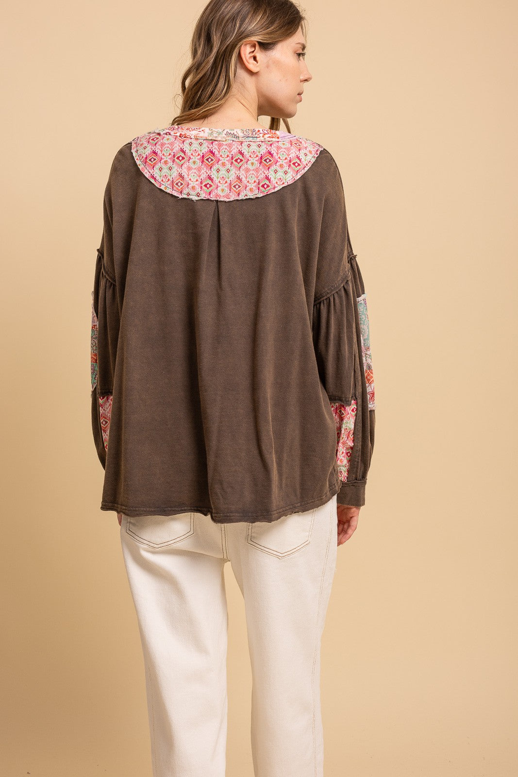 A woman shown from the back wearing a brown long-sleeve top with a colorful floral patterned yoke and sleeve patches, paired with off-white pants, against a solid beige background.