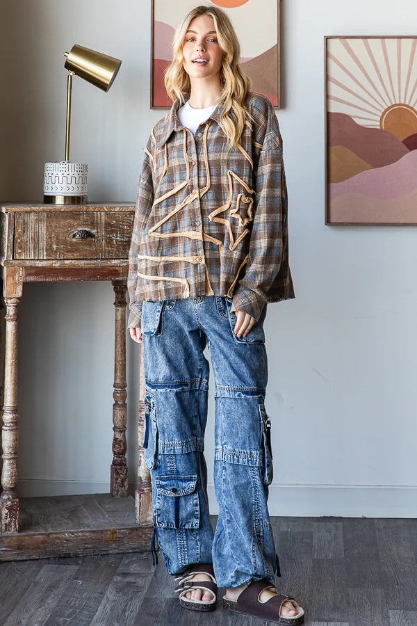 A woman stands indoors wearing a brown plaid button-up shirt with a large star applique on the front, paired with wide-leg blue cargo jeans and brown sandals. The room has a wooden side table with a gold lamp, framed wall art, and a gray wall.
