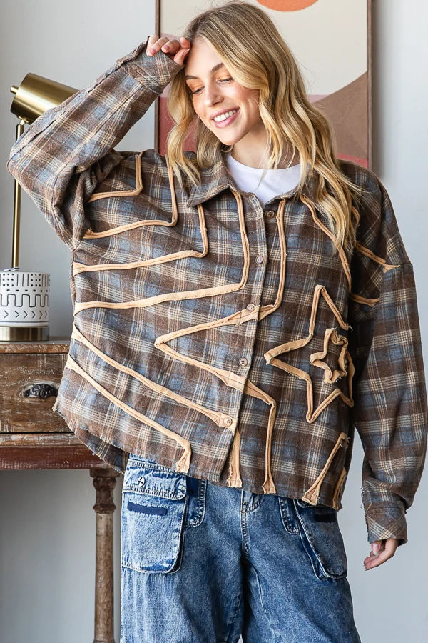 A woman wearing a brown and beige plaid button-up shirt jacket with quilted star and zigzag appliques, styled with blue jeans in an indoor setting.