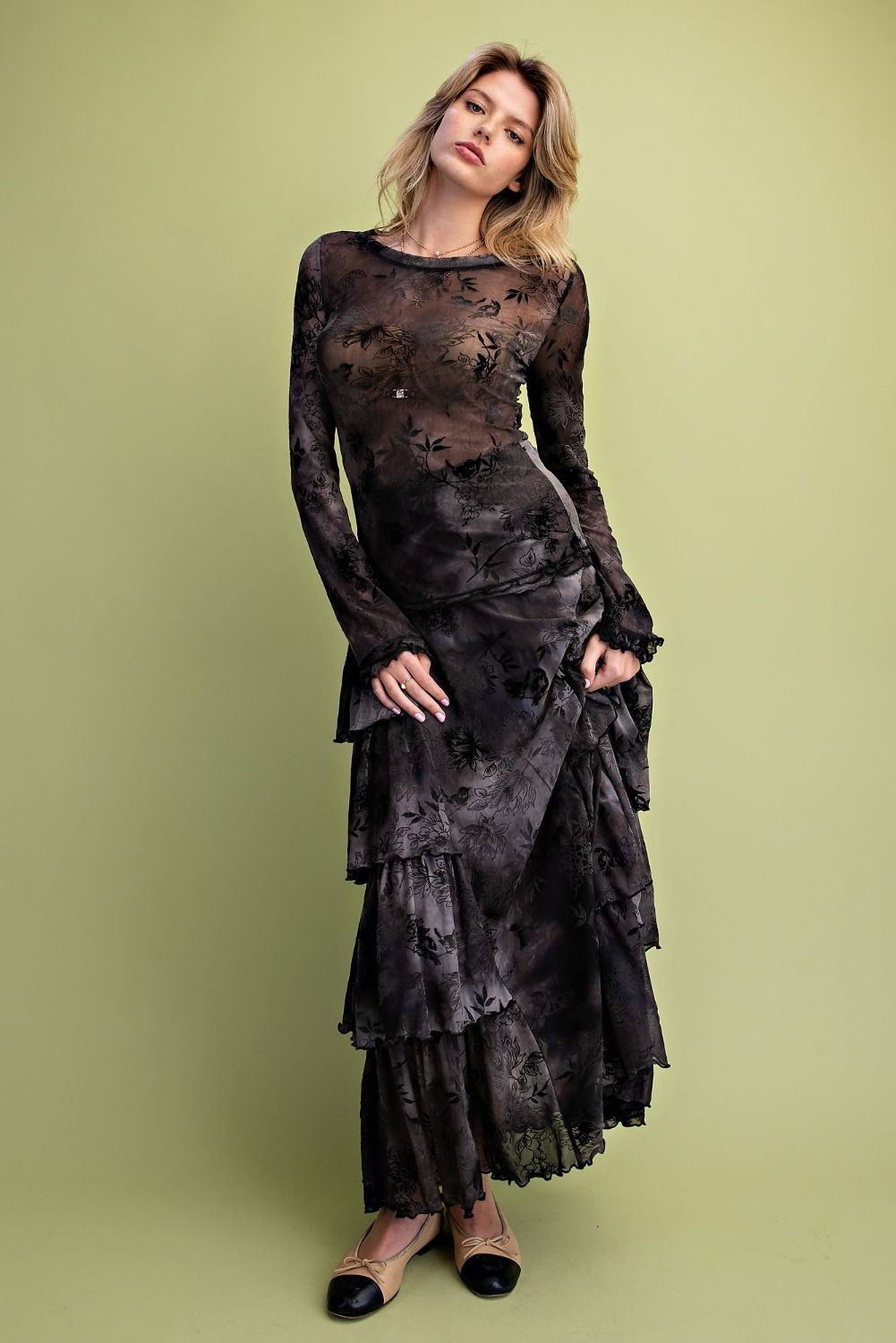 A woman modeling a long-sleeve black sheer mesh dress with a dark floral pattern, featuring a tiered wrap skirt, against a solid olive green studio background.