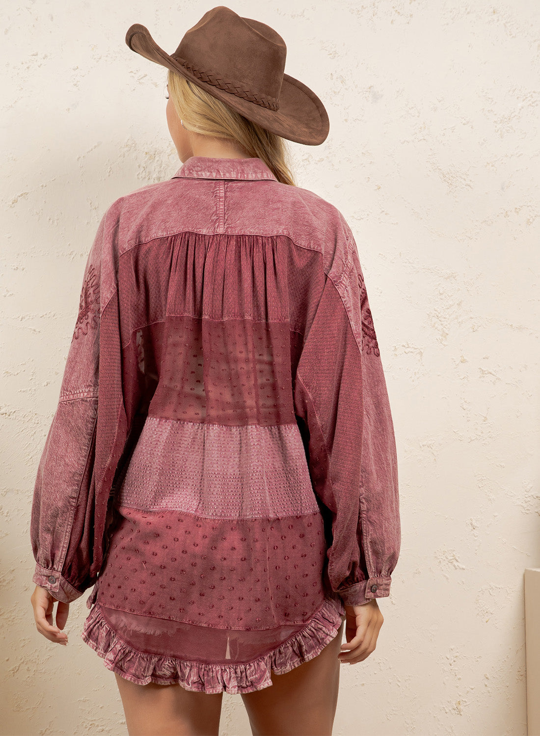 A woman viewed from the back wearing a dusty rose patchwork blouse with long balloon sleeves and a ruffled hem, paired with a brown cowboy hat against a textured off-white wall.