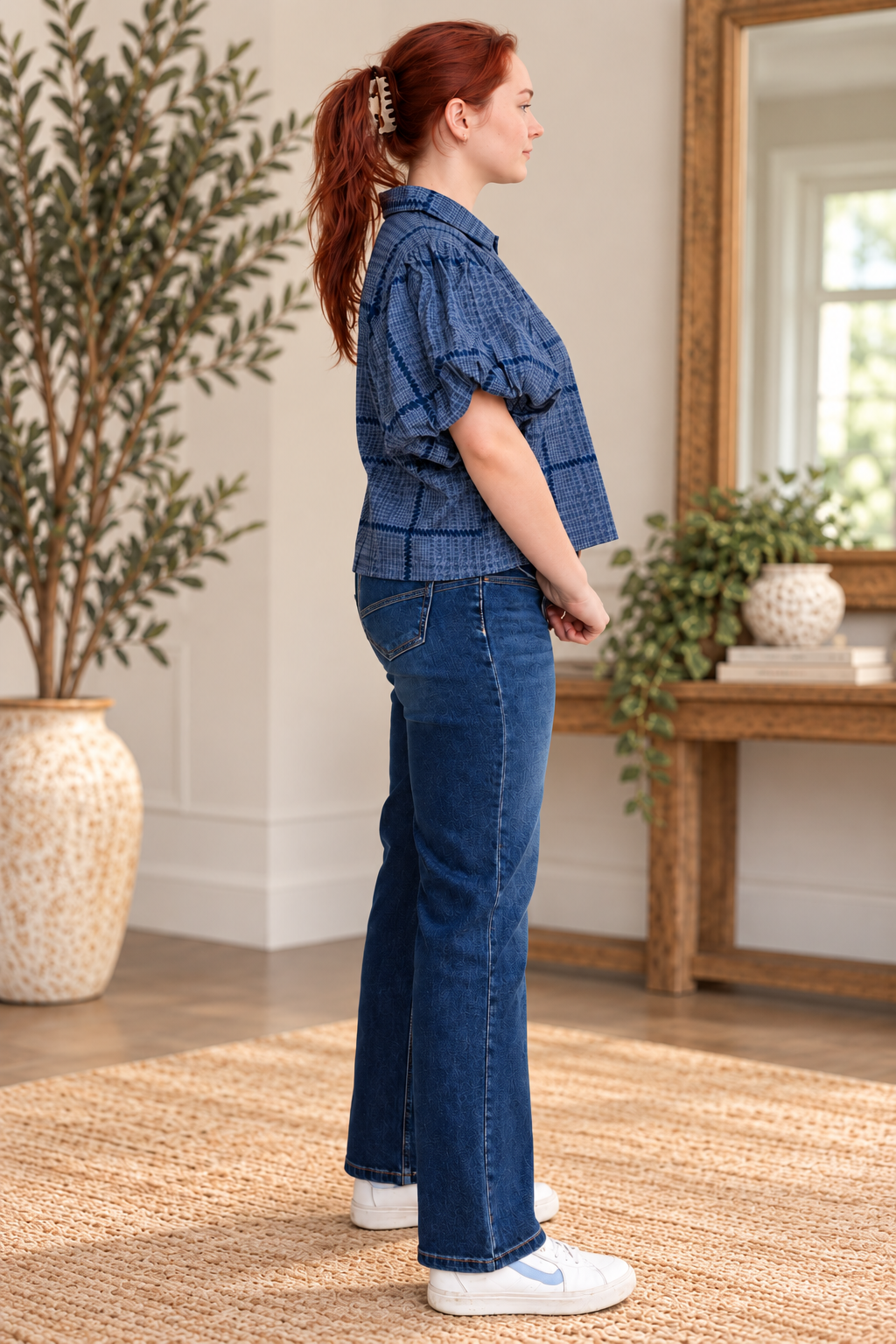 A woman stands in profile wearing a blue plaid short-sleeve cropped top and dark blue flare jeans, paired with white sneakers, in a bright, modern room with a large potted plant and a wooden console table.
