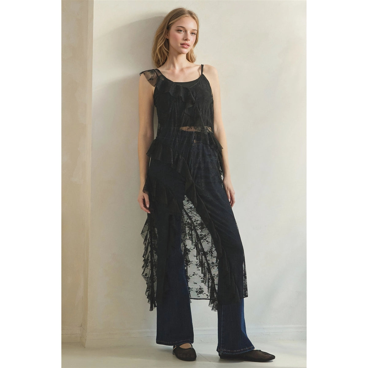 A woman models a black sleeveless lace duster with dark wide-leg jeans against a plain beige wall.