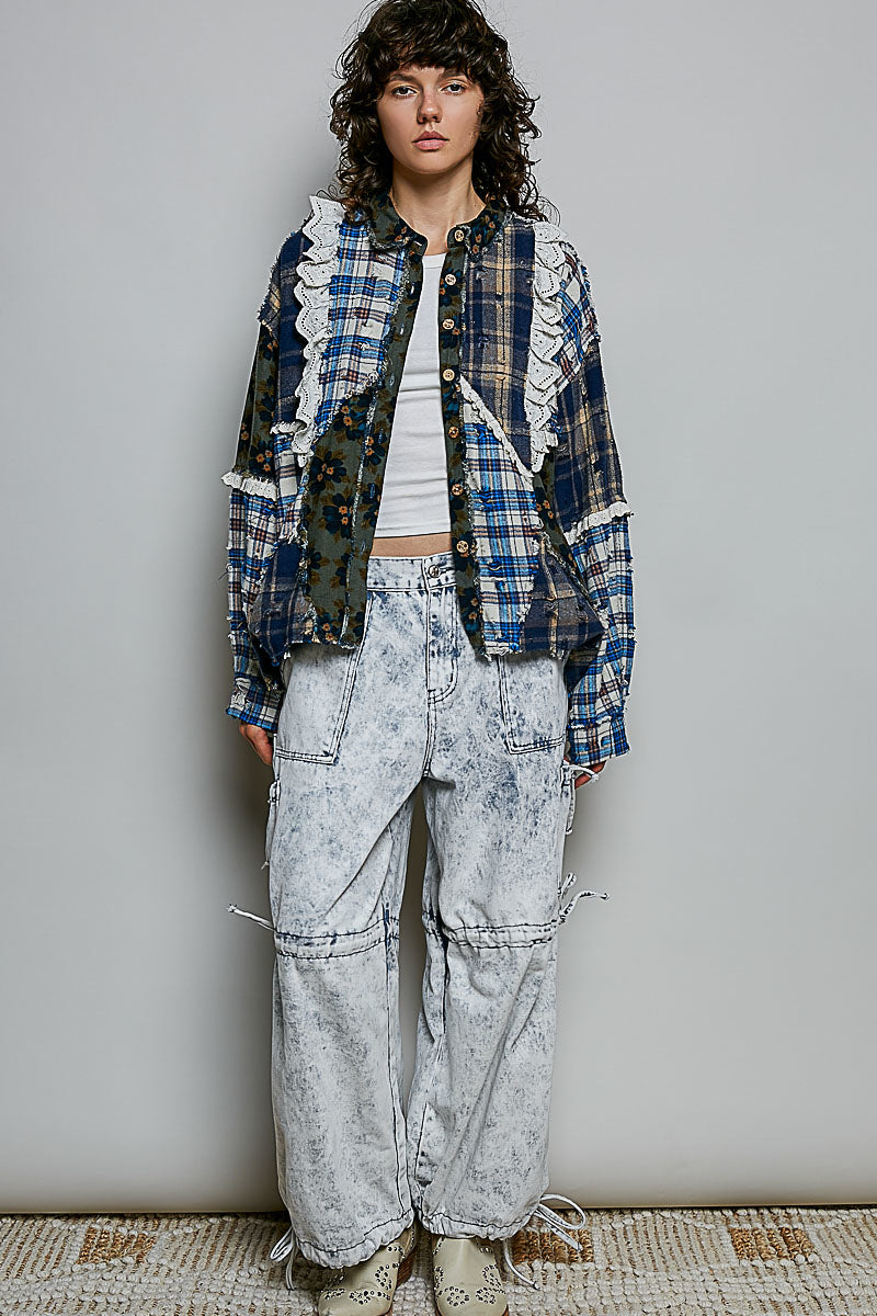 A model stands against a light gray wall wearing a layered outfit featuring a blue and white plaid button-up shirt with ruffle trim over a white crop top, paired with light gray acid-wash wide-leg jeans and beige boots.