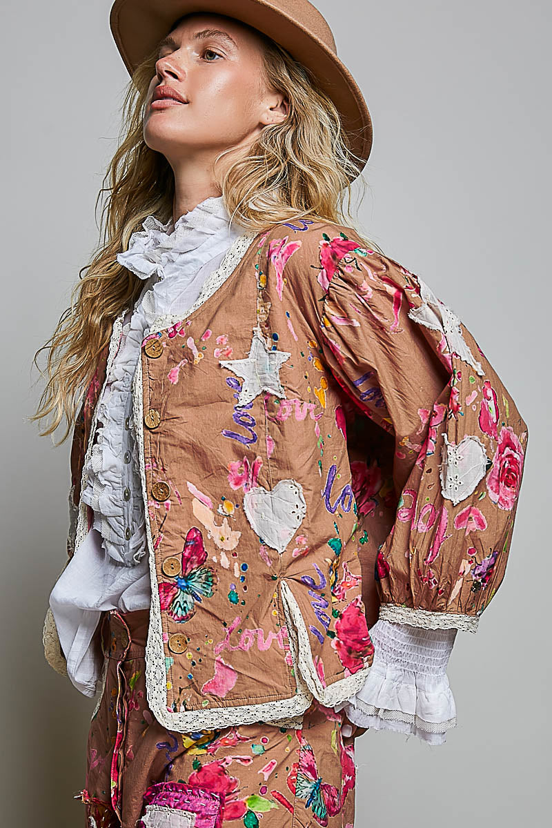 A woman wearing a brown bohemian-style jacket with colorful floral embroidery, white ruffled lace trim, and a matching hat, photographed against a neutral gray background.