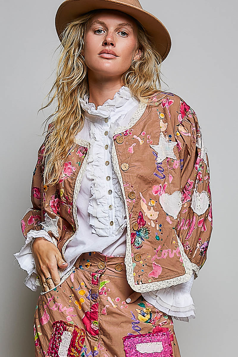 A woman wearing a brown wide-brim hat, a white ruffled blouse, and a brown patchwork jacket with floral and heart appliques, paired with matching patchwork trousers, against a light gray studio background.