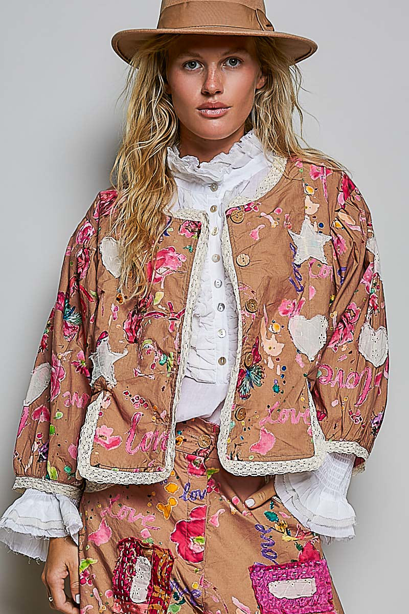 A woman wearing a tan patchwork jacket with pink, white, and multicolor floral embroidery, a matching patchwork skirt, a white ruffled blouse, and a brown wide-brim hat, photographed against a light gray studio background.
