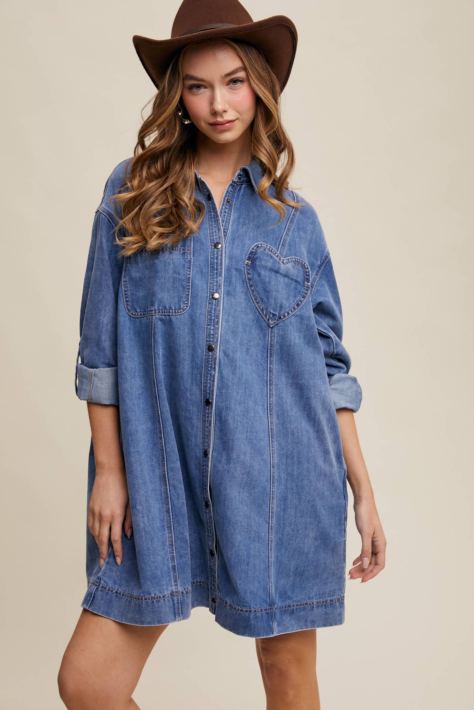 Medium-wash denim shirt dress with snap button front, heart-shaped chest pocket, rolled long sleeves, and a relaxed mini silhouette.