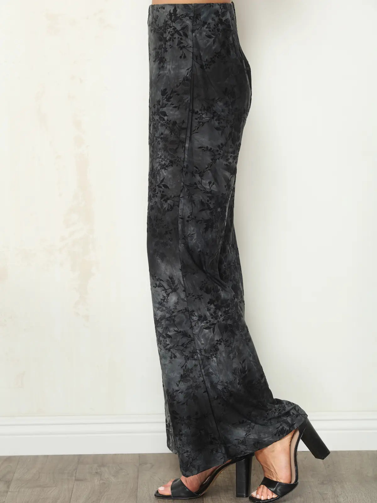 Black floral jacquard wide-leg pants shown on a model from the waist down, paired with black heeled sandals against a neutral off-white wall.