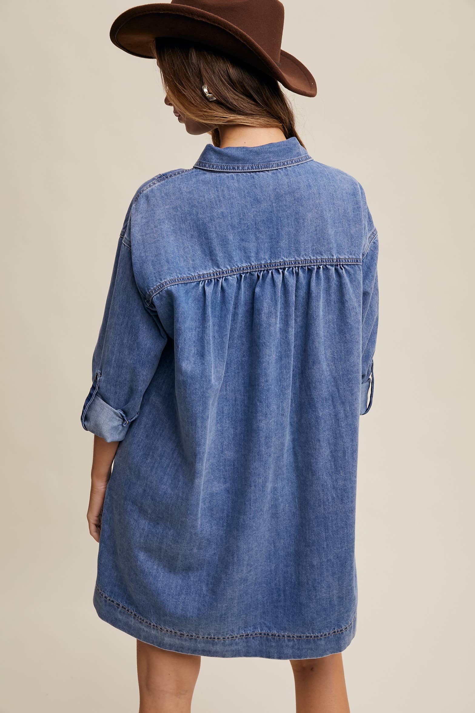 Back view- Medium-wash denim shirt dress with snap button front, heart-shaped chest pocket, rolled long sleeves, and a relaxed mini silhouette.