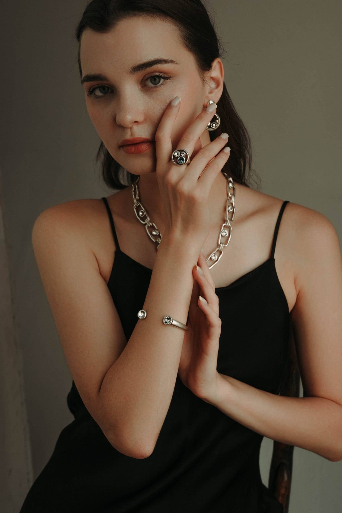 A woman wearing a chunky silver chain necklace, matching silver hoop earrings, a silver ring, and a silver cuff bracelet, posing in a black spaghetti-strap dress against a dark gray background.