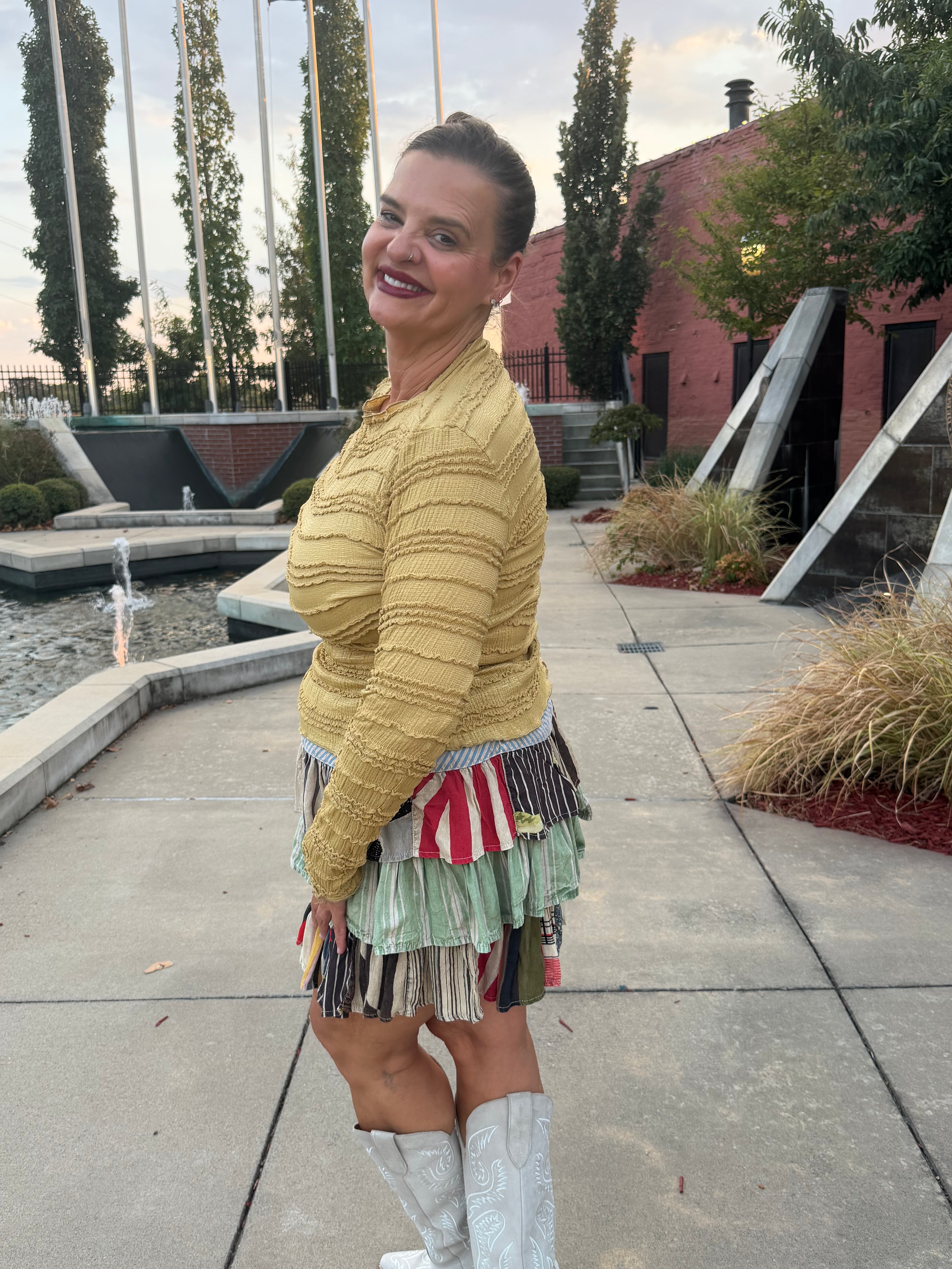 A woman wearing a textured mustard yellow knit sweater and a colorful striped fringe skirt, posing outdoors on a concrete walkway with a modern building and trees in the background.