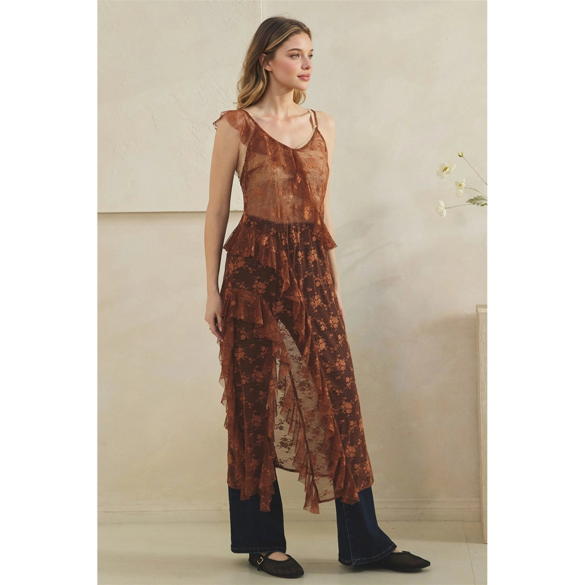 A woman models a copper sheer floral maxi duster with a ruffled, tiered overlay, paired with dark wide-leg jeans and black flat shoes, standing against a neutral beige wall.