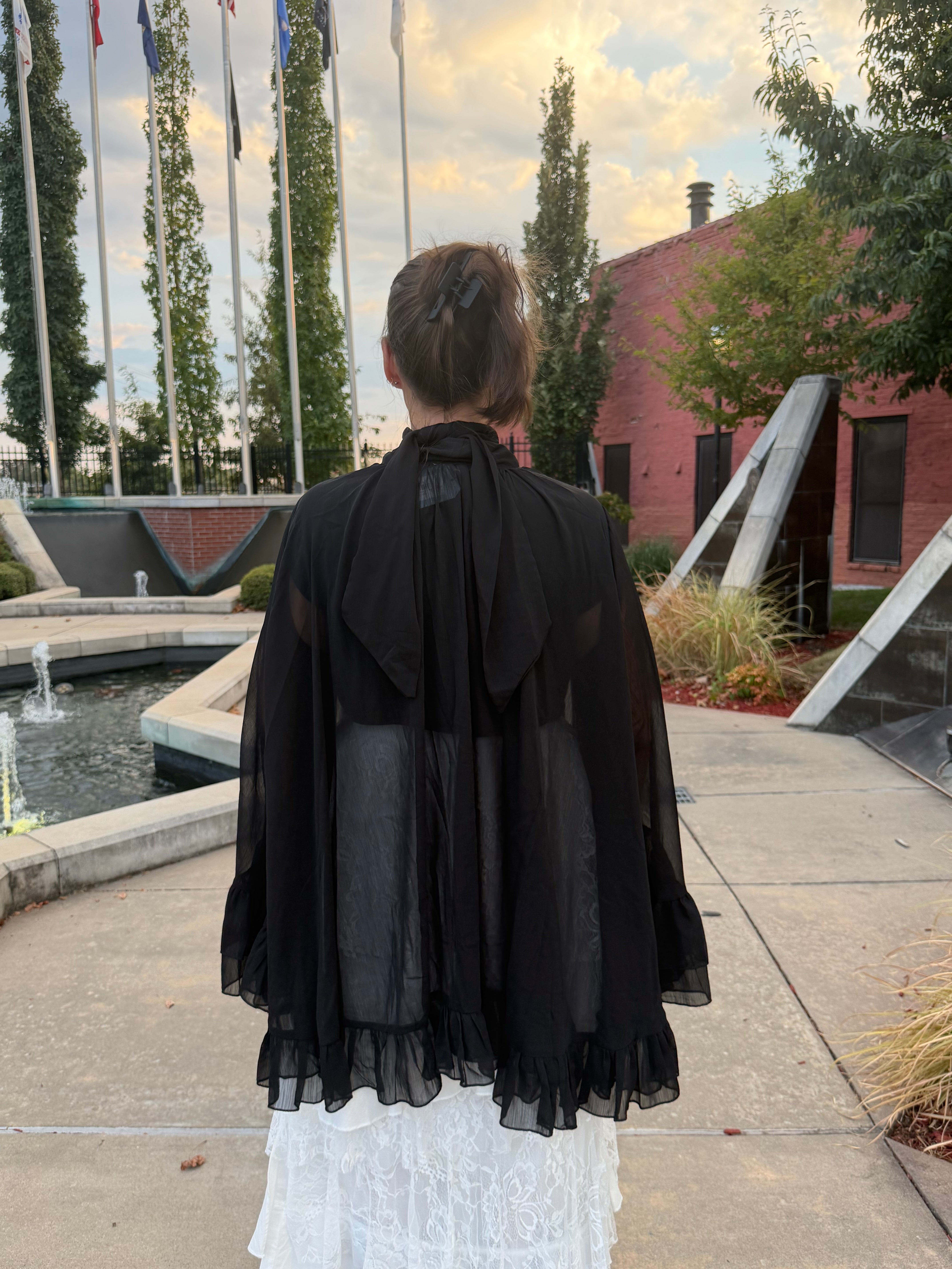A woman viewed from behind wearing a sheer black long-sleeve cape-style top with a large bow at the back, standing outdoors on a paved walkway near a fountain and brick building.