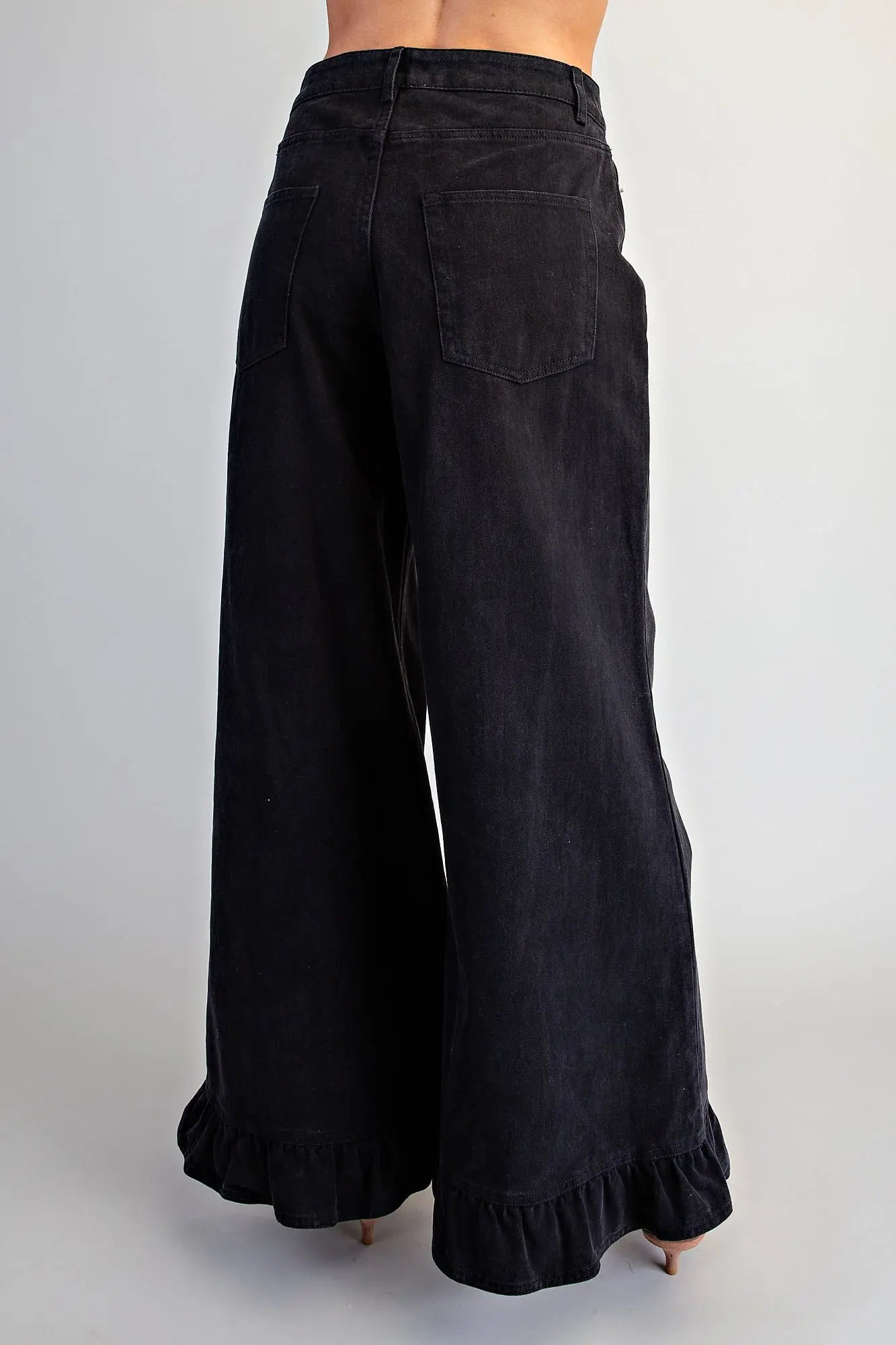 Back view of black high-rise wide-leg denim pants with single back patch pocket and ruffled hem, styled with nude kitten heel mules 