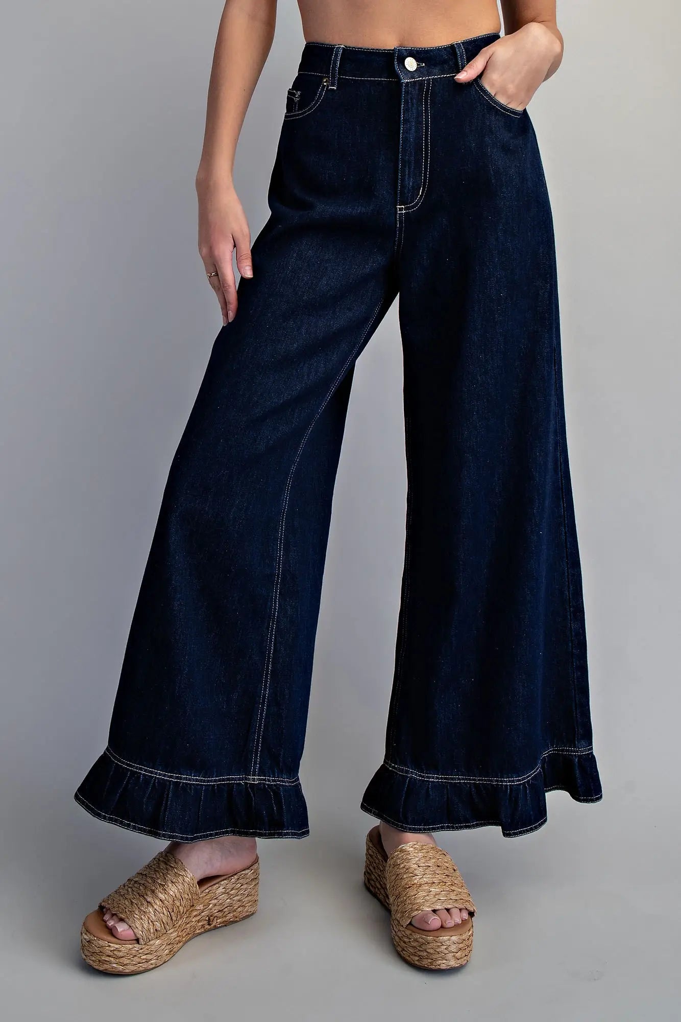Front view of women's dark wash high-rise wide-leg denim pants with contrast white stitching and ruffled hem, styled with woven platform wedge sandals