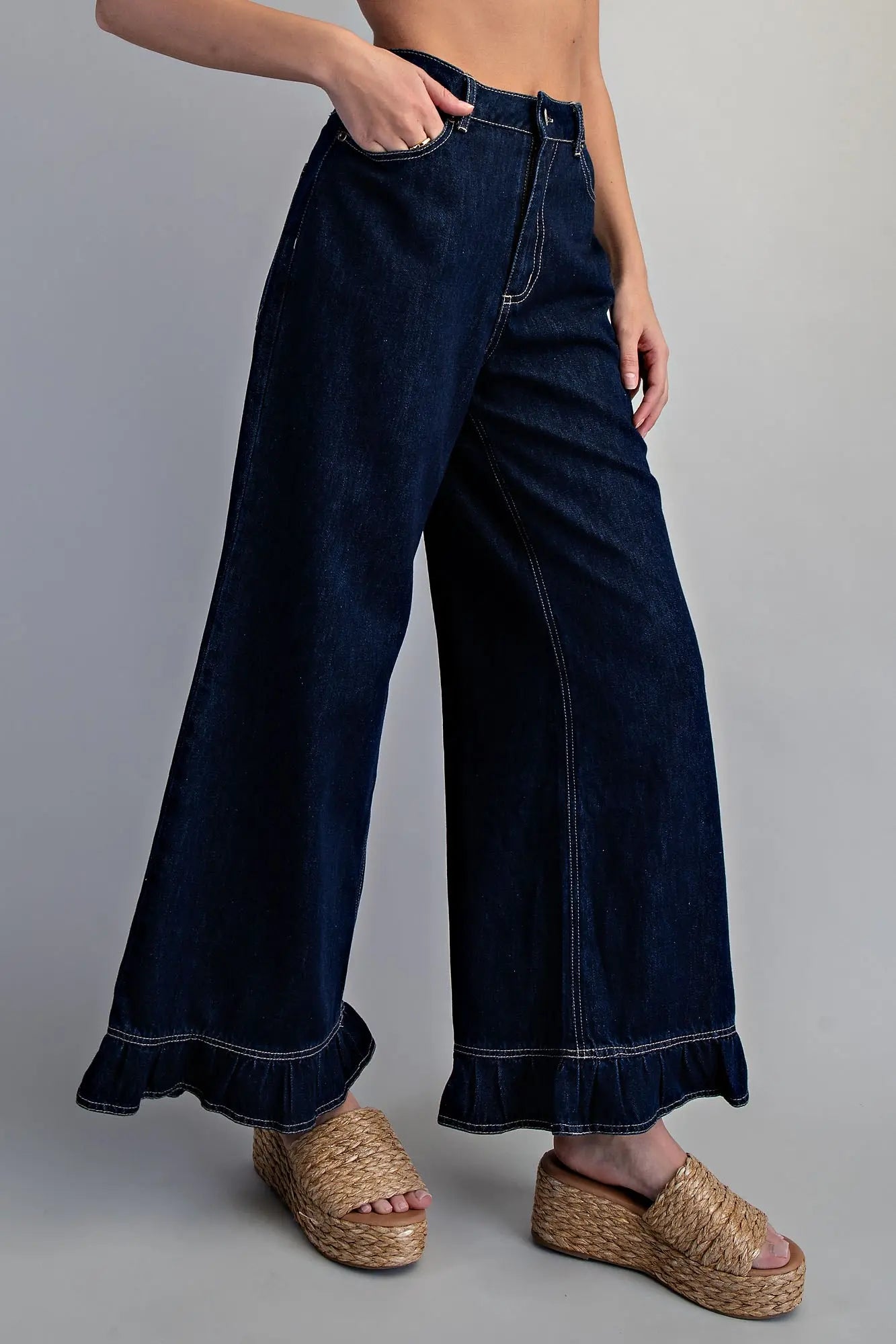 Side view of women's dark wash wide-leg denim pants showing dramatic flare silhouette, contrast stitching, and ruffled hem detail, paired with woven wedge sandals 