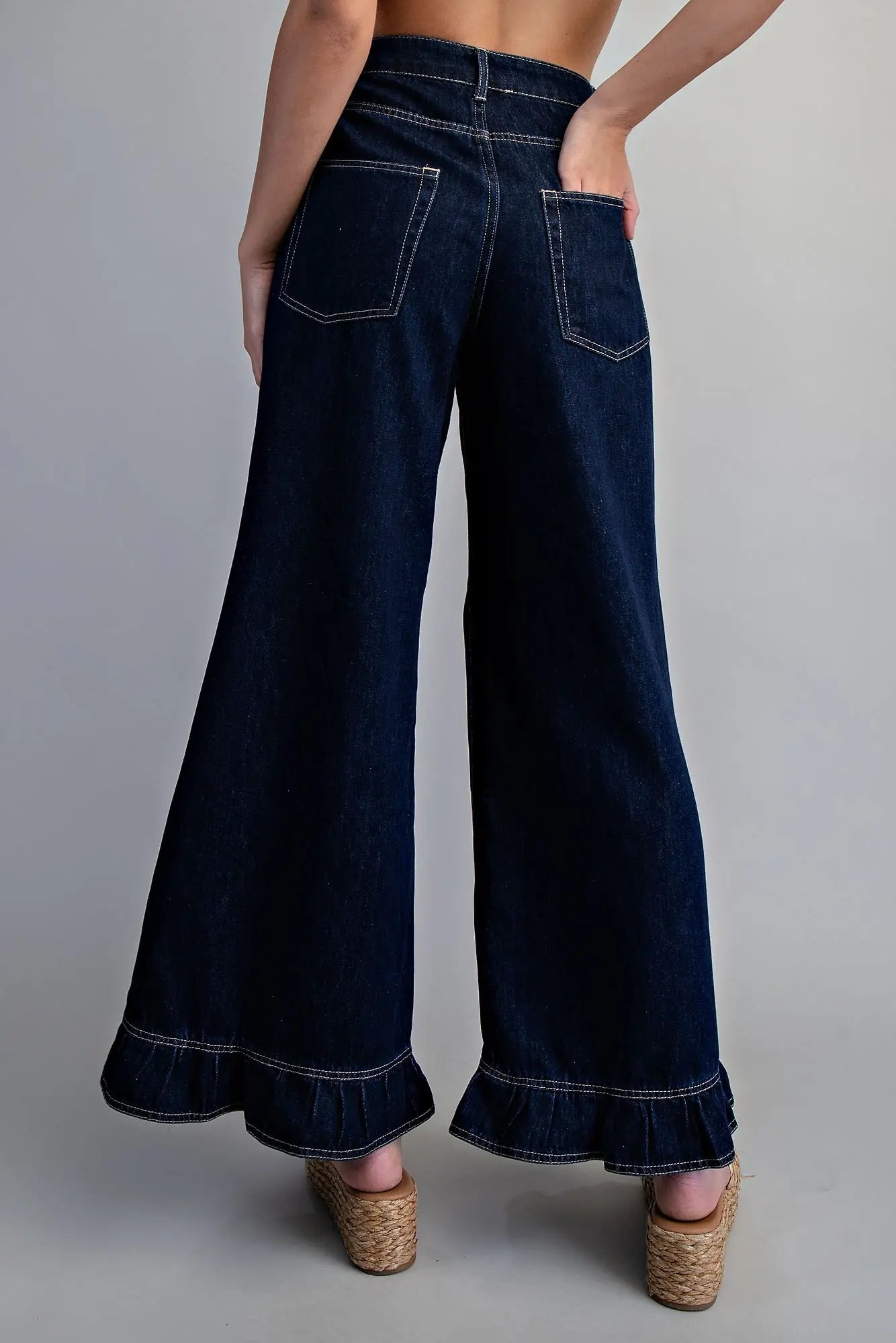 Back view of dark wash wide-leg denim pants with contrast white stitching, back patch pockets, and ruffled hem, styled with woven platform wedge sandals