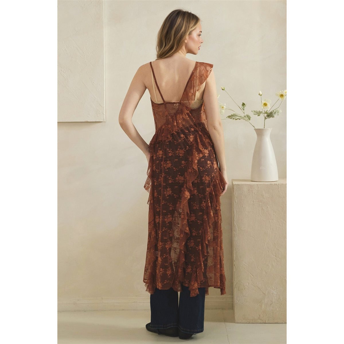 A woman stands with her back to the camera wearing a copper sheer floral duster with a low open back, paired with dark wide-leg pants, in a warm beige room next to a white vase with flowers.