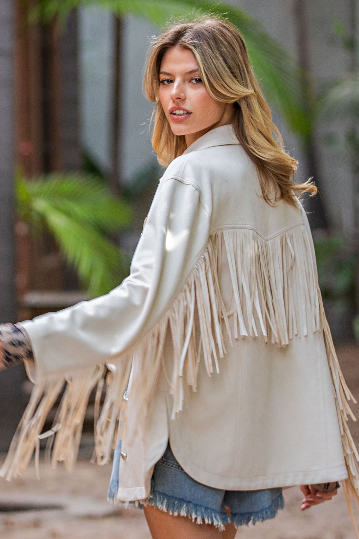 A woman wearing a cream-colored fringe jacket and denim shorts, photographed outdoors with a blurred background of greenery and stone walls.