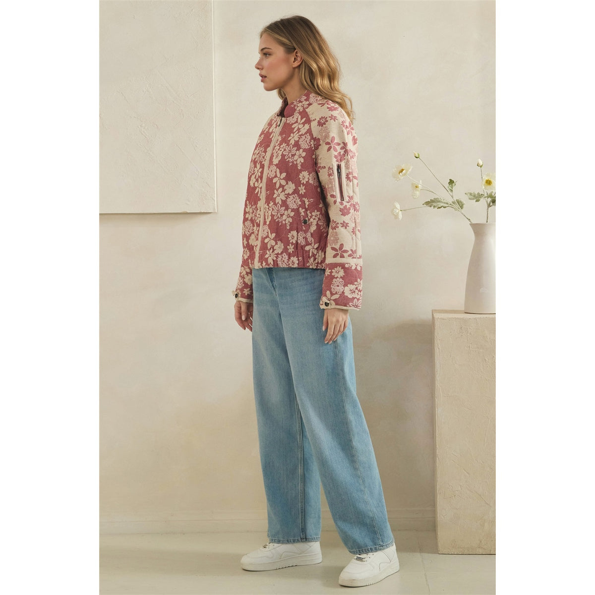 A woman stands in profile wearing a pink and beige floral jacket, wide-leg light blue jeans, and white sneakers in a minimalist beige interior with a vase of flowers.