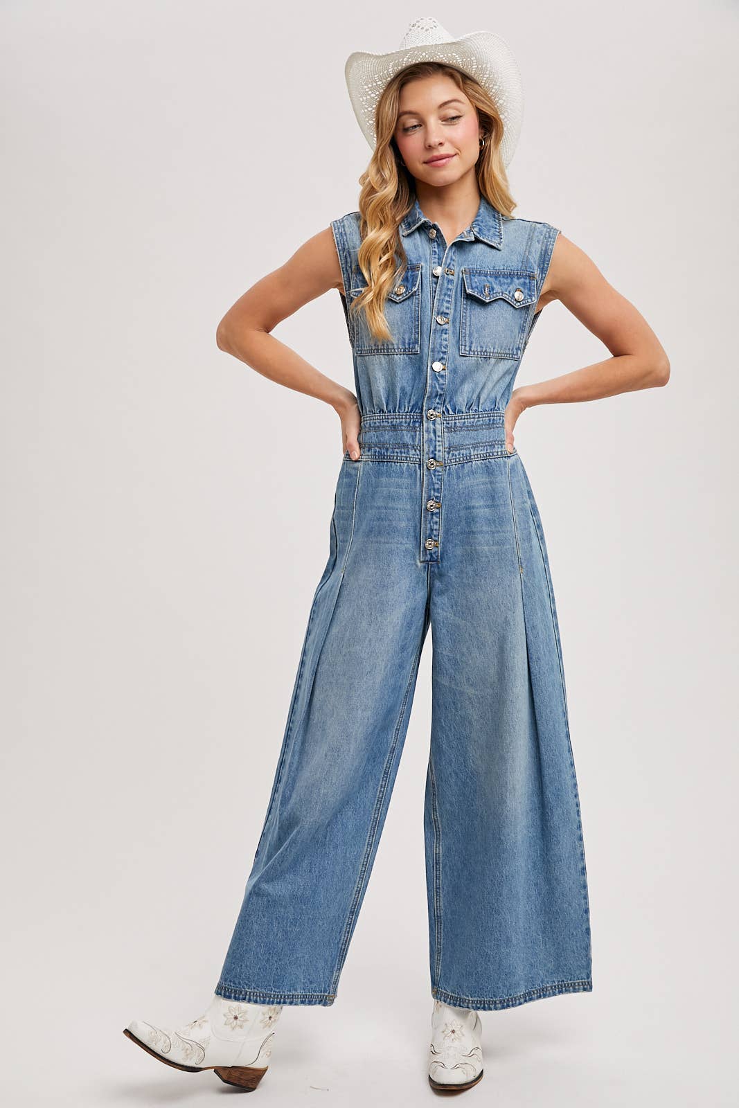 Sleeveless denim jumpsuit with collared button-front, chest flap pockets, and wide-leg palazzo silhouette, shown on model with slide sandals.