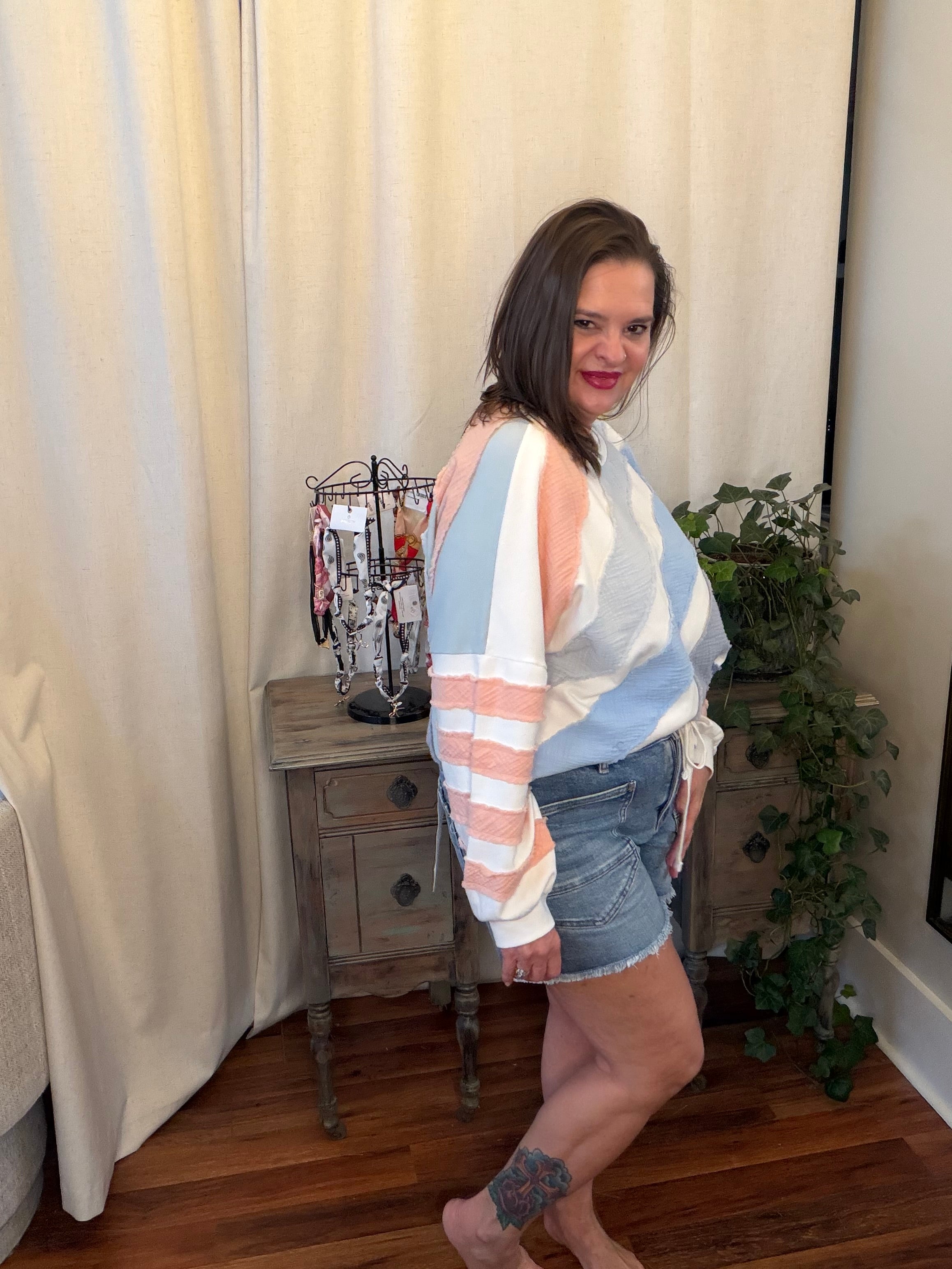 Woman wearing a striped shirt and denim shorts standing in a room with curtains and a plant.
