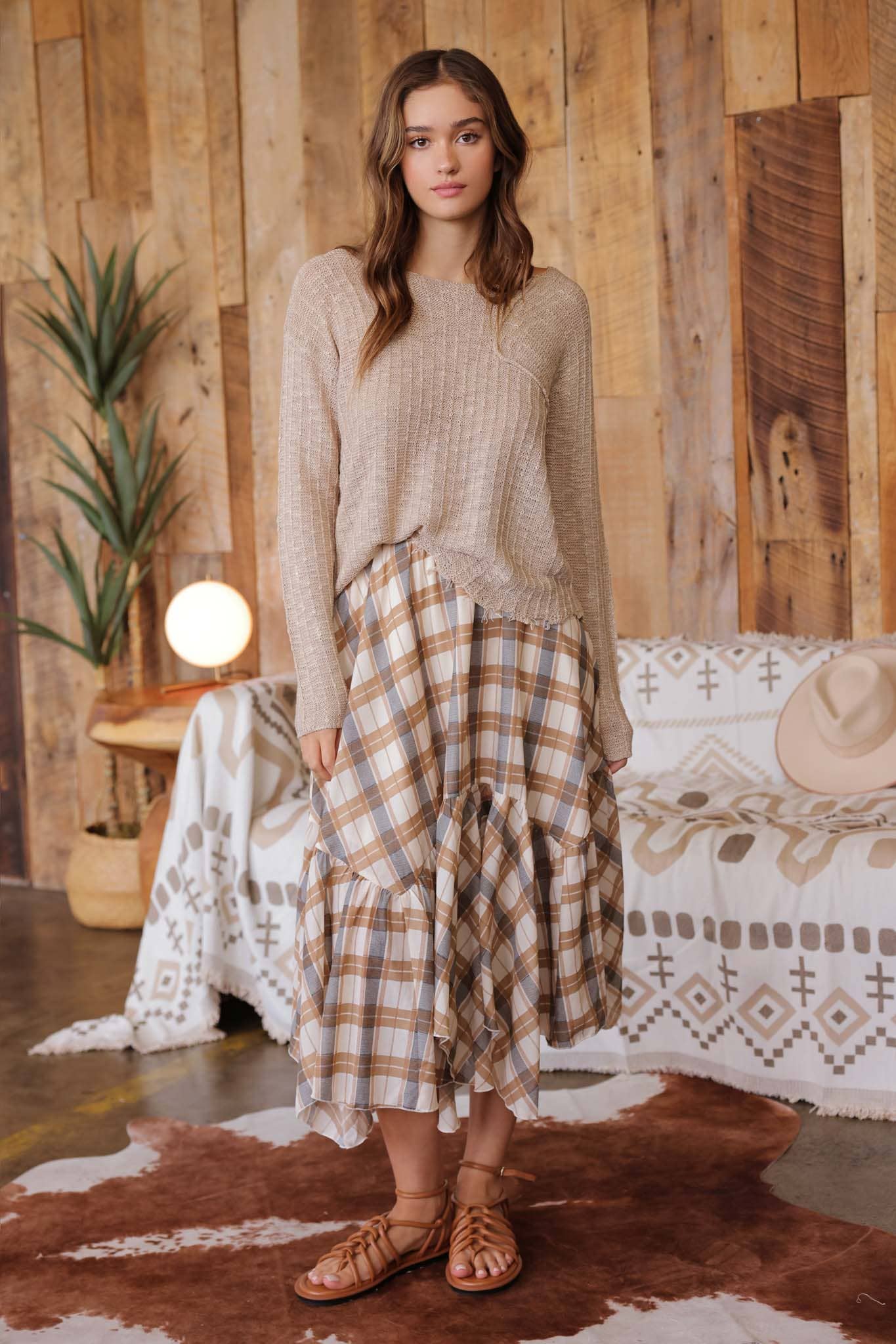 Front view of model wearing taupe distressed lightweight sweater with ribbed knit texture, raw edge hem, and relaxed fit paired with tan plaid ruffle skirt
