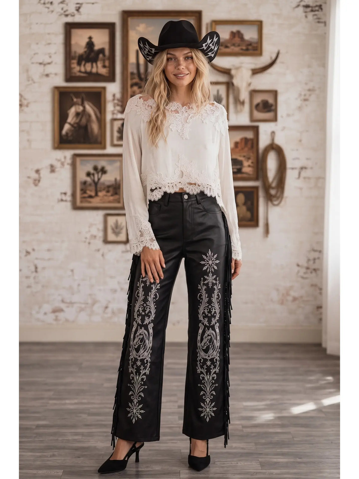A woman stands in a rustic indoor setting wearing a white lace-trim blouse, black fringed western-style pants with silver embroidery, a black cowboy hat, and black heels.