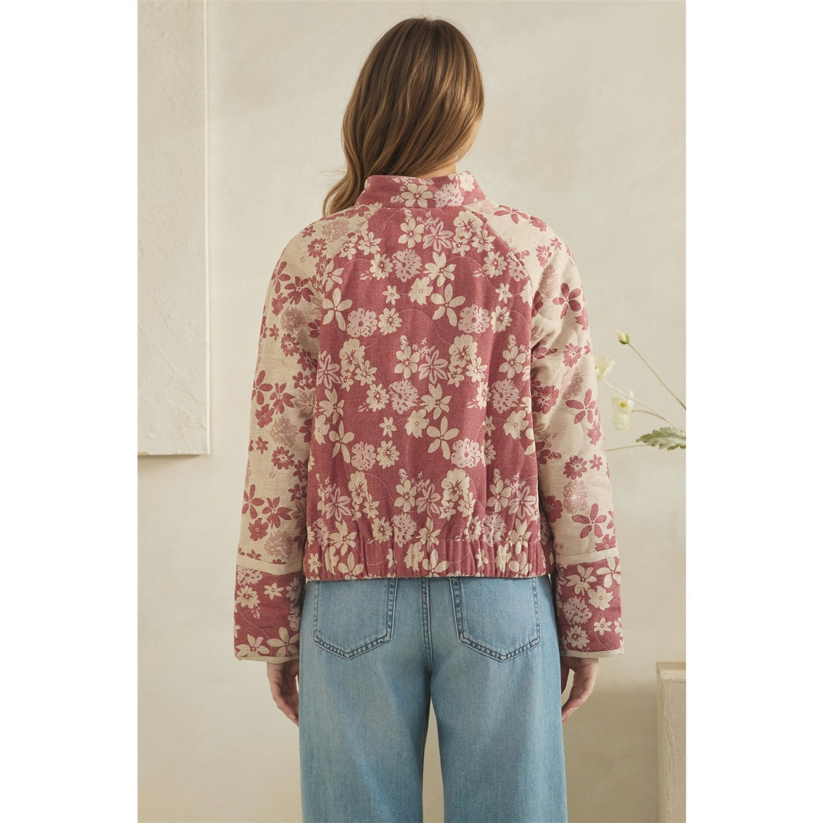 A woman viewed from behind wearing a cropped floral jacket with a dusty rose and cream pattern, paired with light blue jeans, in a softly lit indoor setting.