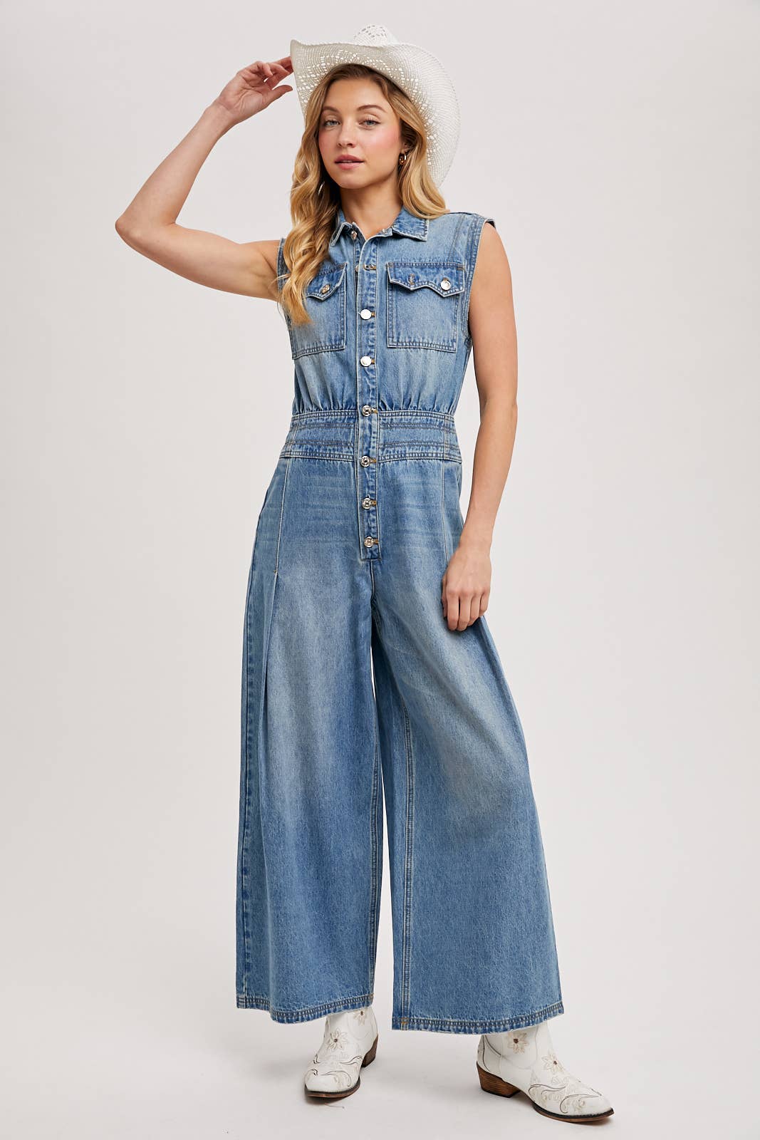 Sleeveless denim jumpsuit with collared button-front, chest flap pockets, and wide-leg palazzo silhouette, shown on model with slide sandals.