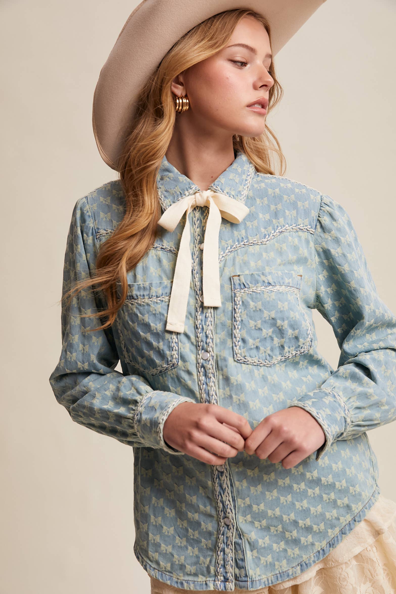 Light-wash denim blouse with cream bow-tie collar, tonal mini-bow pattern, braided contrast stitching, snap front, and two chest pockets.