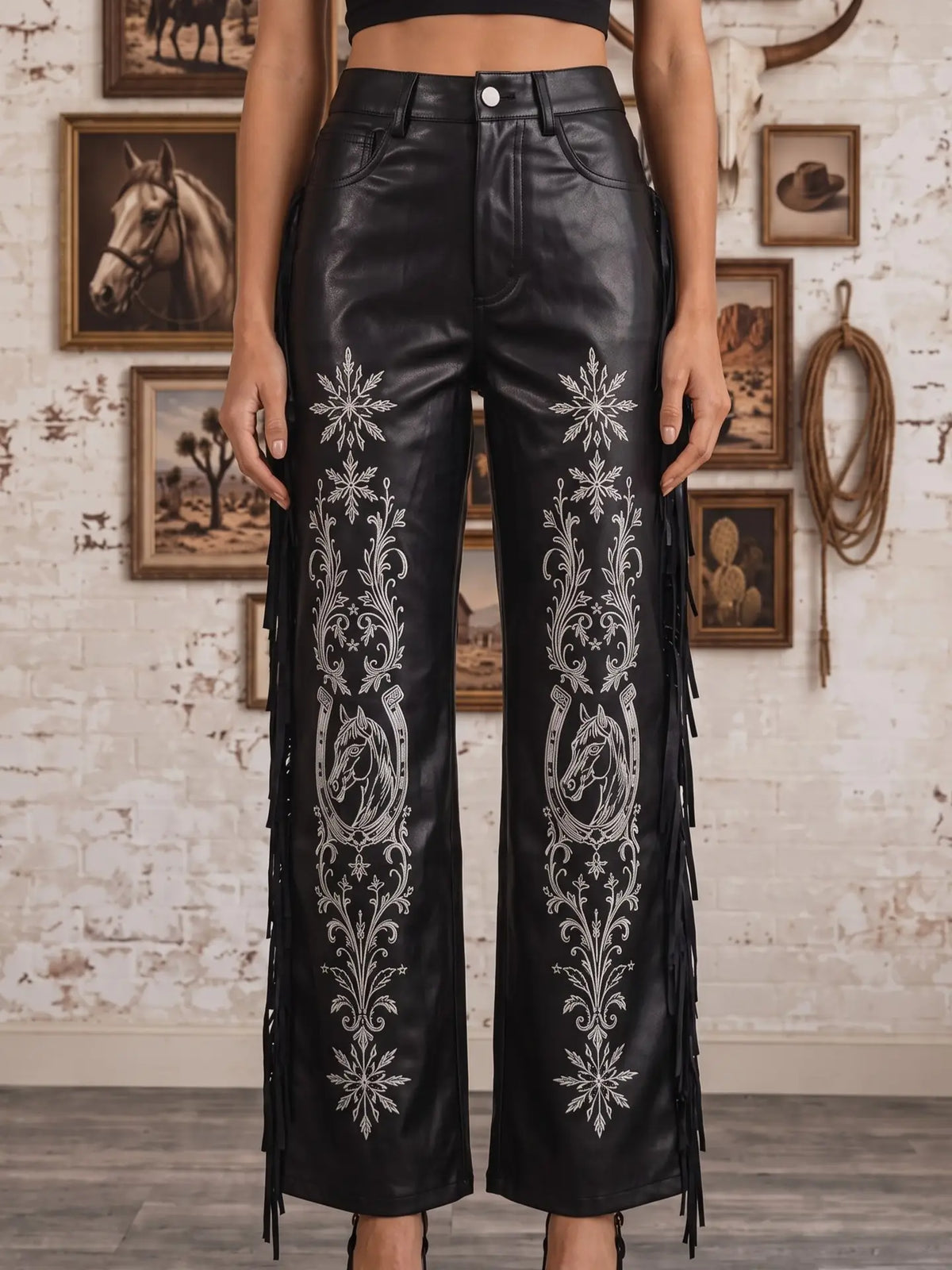 Black faux leather wide-leg pants with silver western embroidery and fringe details, shown on a model in a rustic indoor setting.