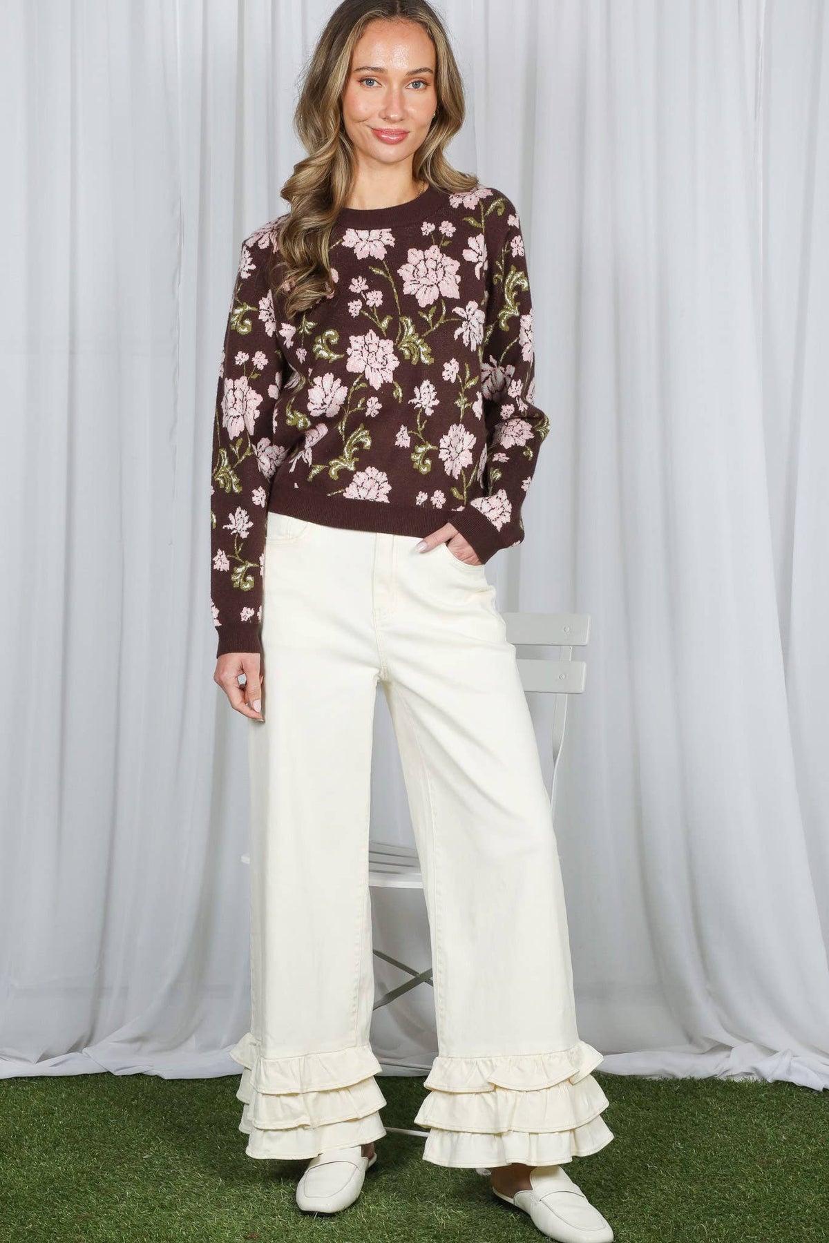 A woman modeling a dark brown floral knit sweater and cream wide-leg ruffled pants, standing against a white draped backdrop with artificial green turf at the bottom.