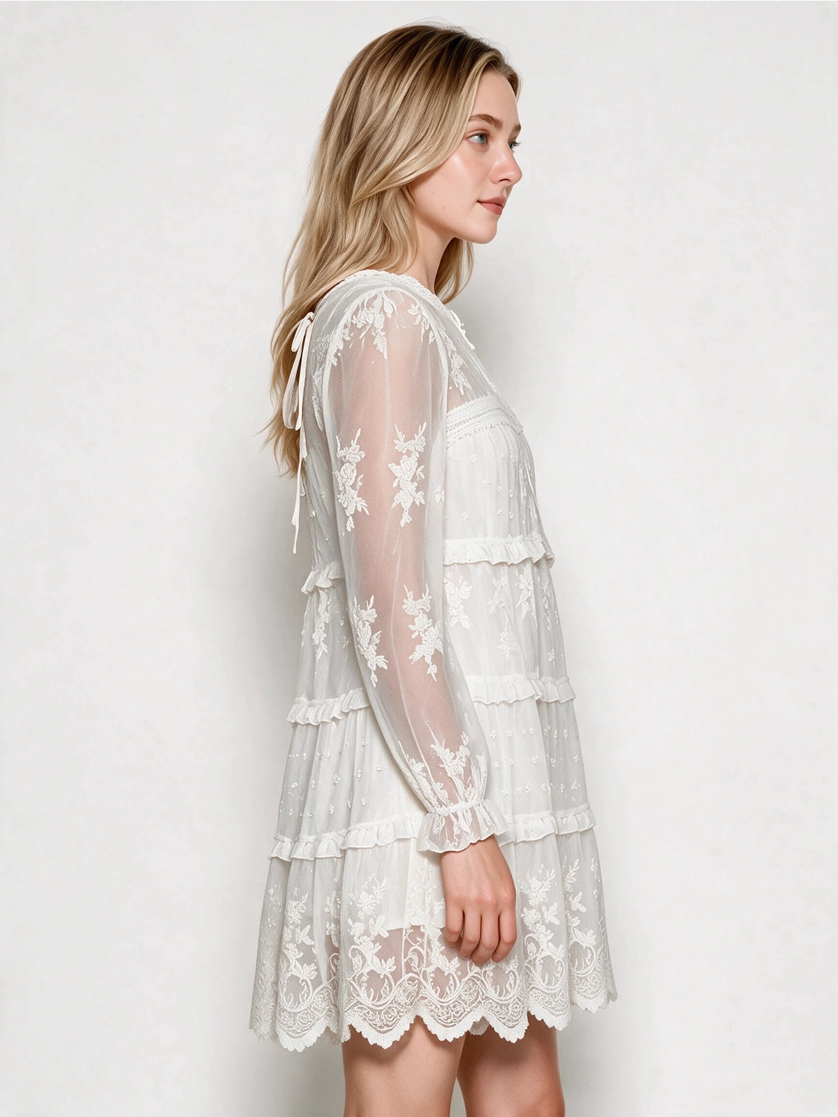 Side view of the white tiered lace dress showing sheer floral embroidered sleeves, ruffled cuffs, and a scalloped hem.
