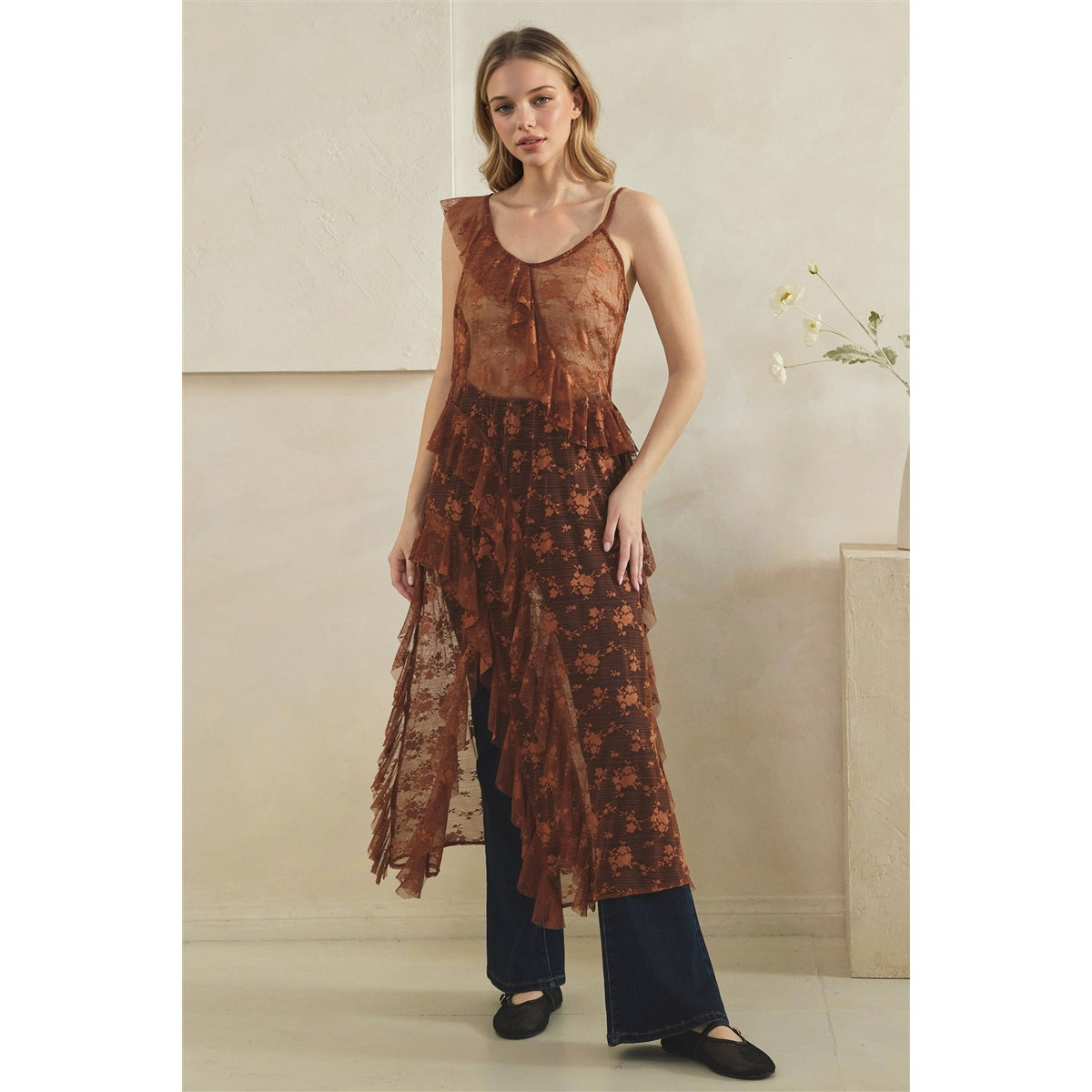 A woman models a copper sheer floral lace tunic with ruffled layers over dark wide-leg pants, standing in a minimalist beige room.