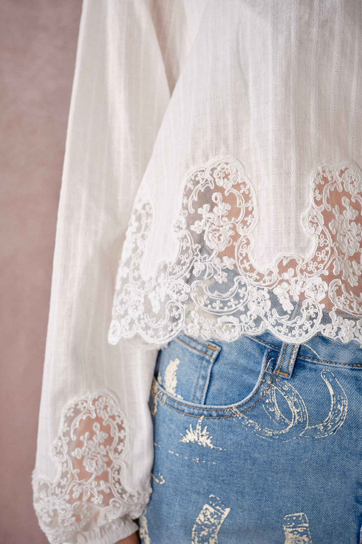 White long-sleeve blouse with intricate floral lace trim at the cuffs and hem, styled with distressed blue jeans against a soft pink background.