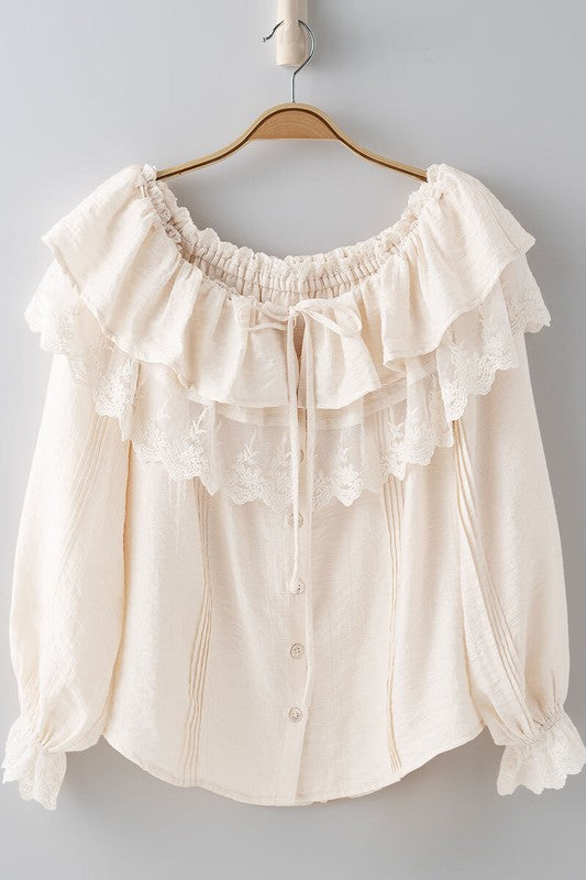 Cream-colored blouse with ruffled details on a hanger against a gray background