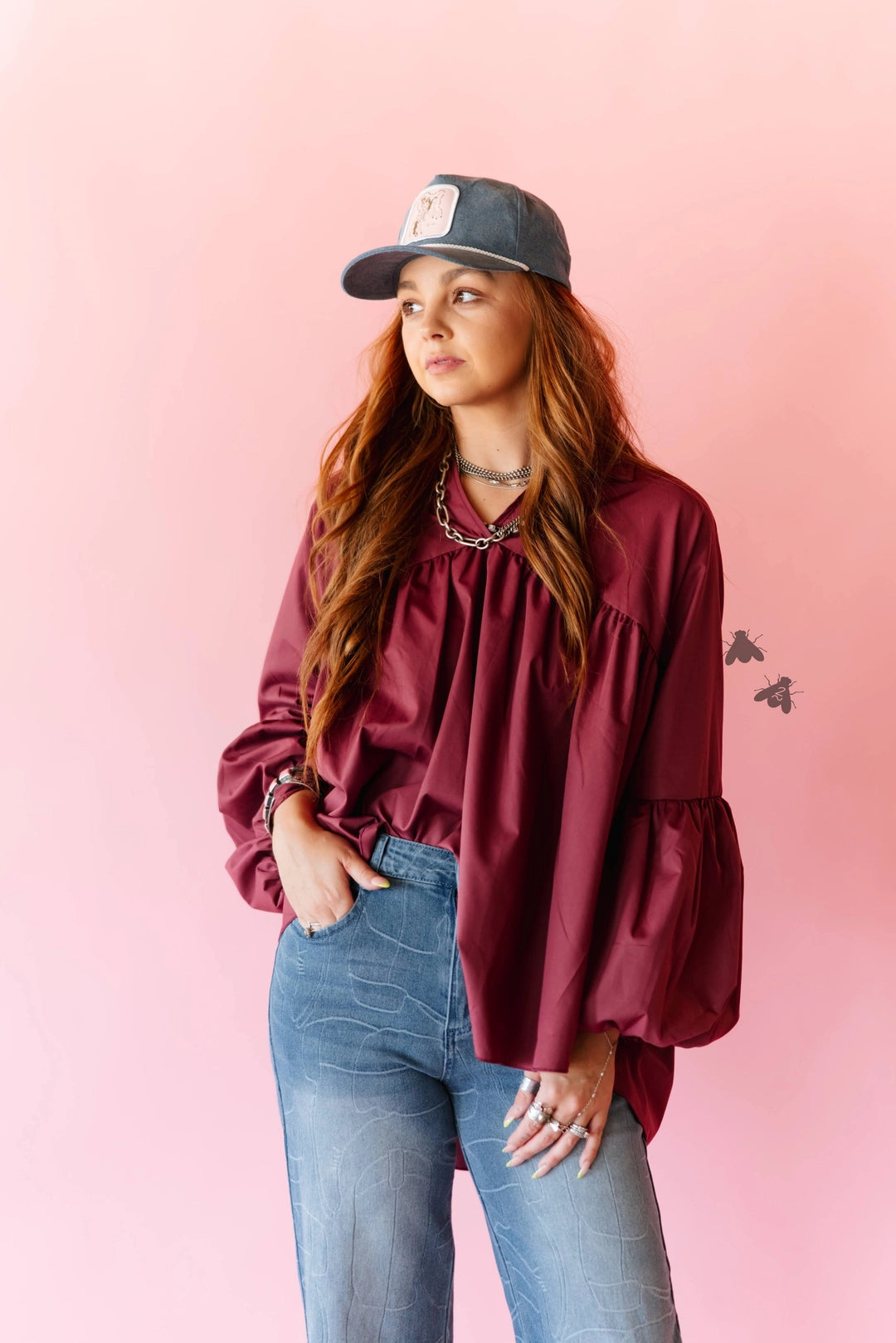 A woman wearing a long-sleeve maroon blouse with a gathered neckline and ruffled sleeves, paired with blue jeans and a dark baseball cap, standing against a solid pink background.