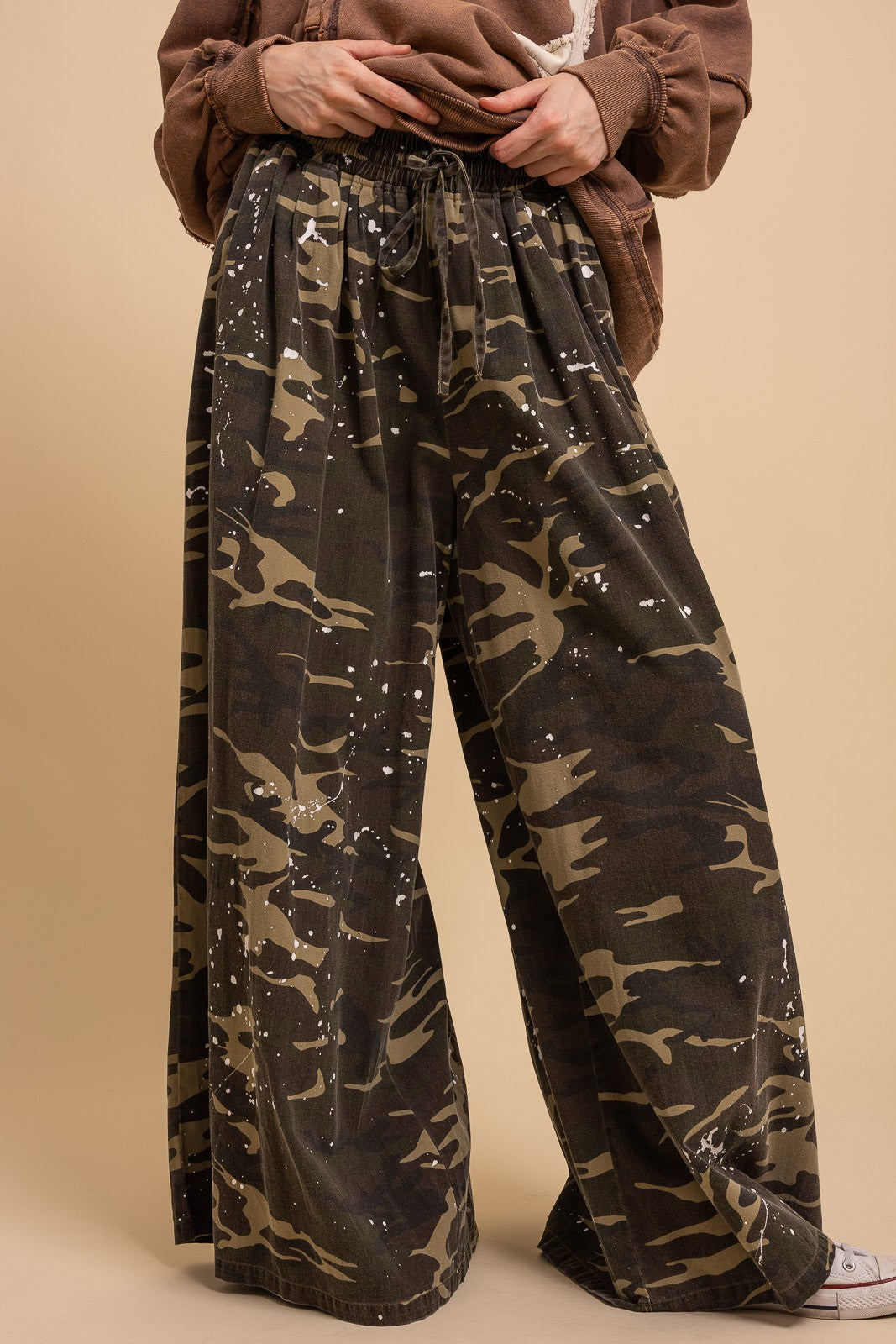 Front close-up of Blue Velvet paint splash camo wide leg pants showing elastic drawstring waistband and white paint splash camo print detail, brown jacket styling
