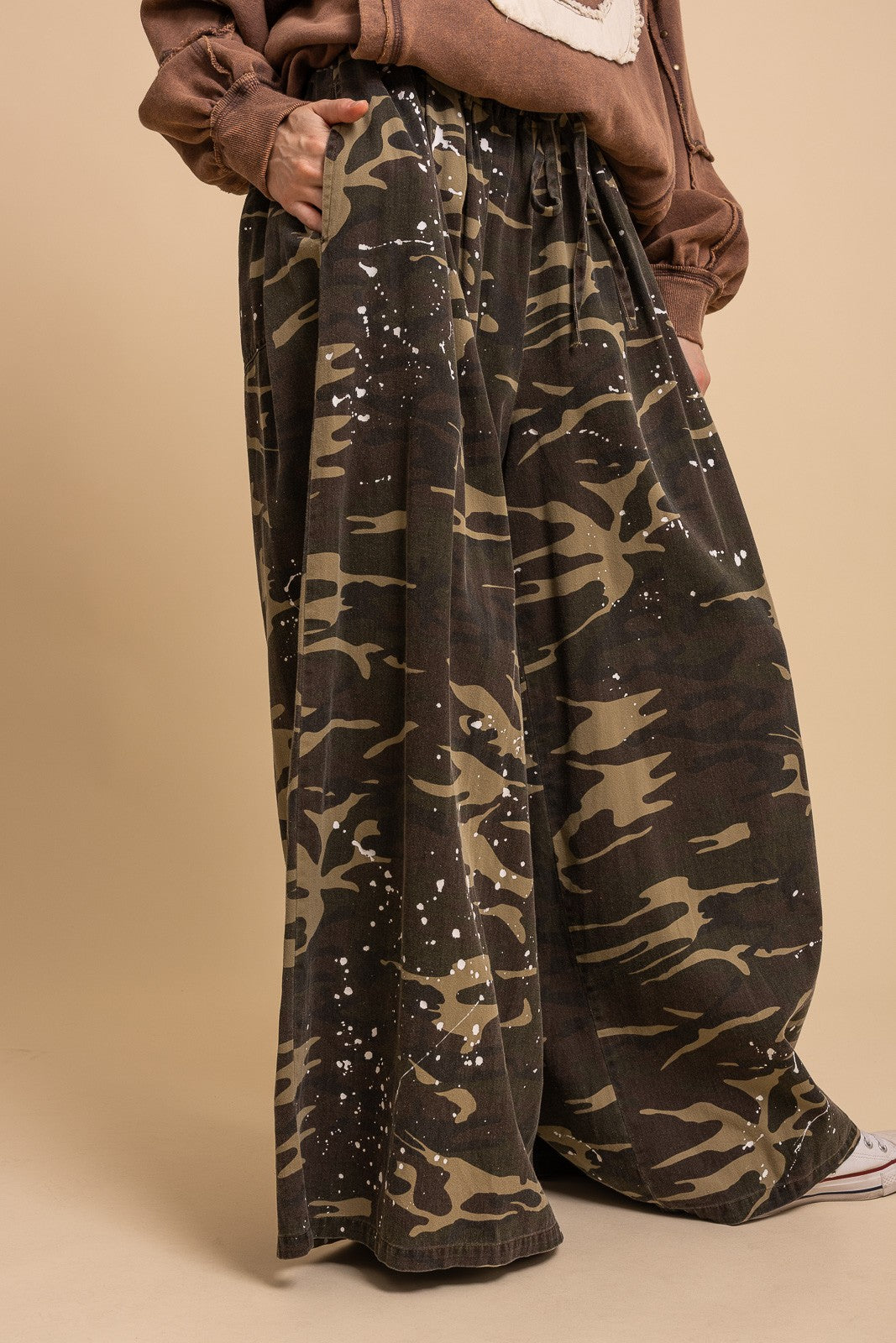Front three-quarter close-up of Blue Velvet paint splash camo wide leg pants showing drawstring waist, paint splash detail, and wide leg drape with brown jacket
