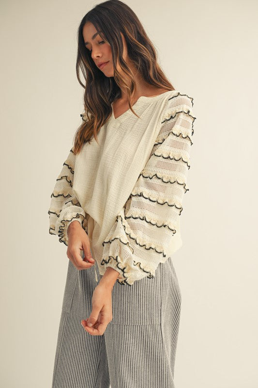 Front view of cream gauze blouse with sheer lace balloon sleeves, black-trimmed scalloped ruffles and a V-neck, styled with gray striped wide-leg pants.