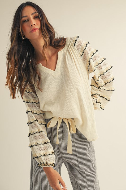 Front view of cream gauze blouse with sheer lace balloon sleeves, black-trimmed scalloped ruffles and a V-neck, styled with gray striped wide-leg pants.