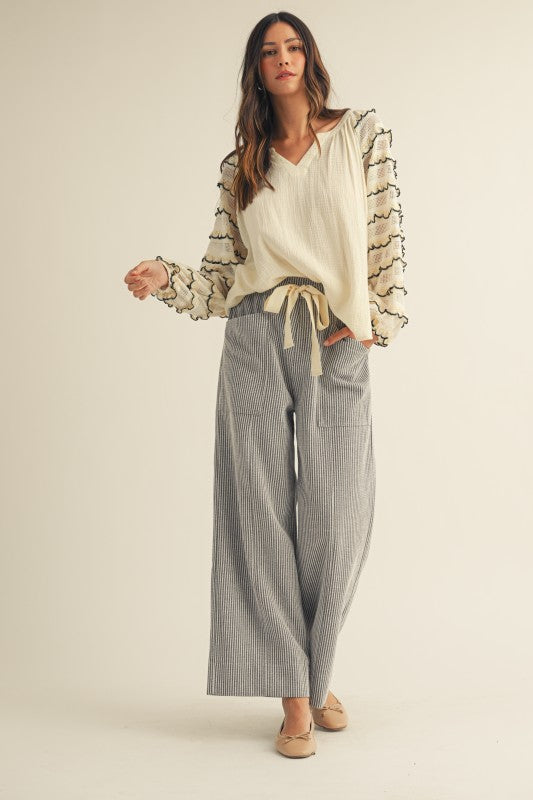 Front view of cream gauze blouse with sheer lace balloon sleeves, black-trimmed scalloped ruffles and a V-neck, styled with gray striped wide-leg pants.